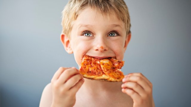 Article thumbnail: Cute little boy eating pizza. The shirtless boy aged five is very happy, is smiling and looking away. There is a gray wall behind the boy.