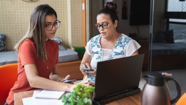 Article thumbnail: Mother and daughter doing finances together at home. They talk and handle some bills, looking and pointing at laptop screen