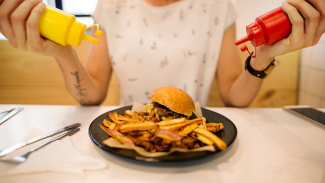 Article thumbnail: Cropped shot of a woman adding mustard and tomato ketchup to the plate with burger and chips at a restaurant